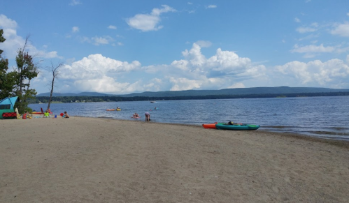 A sandy beach with people enjoying the water, kayaks, and a scenic view of clouds and mountains in the background.