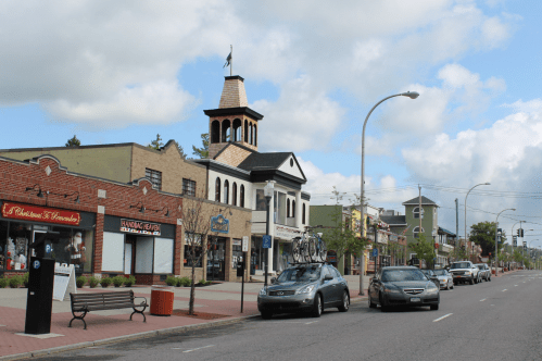 A street view featuring shops, a clock tower, and parked cars under a cloudy sky.