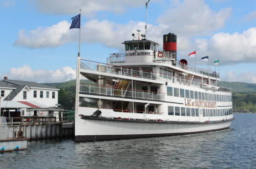 A large white steamboat docked at a pier, with flags flying and mountains in the background under a partly cloudy sky.