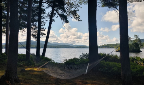 A serene lakeside view with a hammock strung between two trees, surrounded by mountains and fluffy clouds.