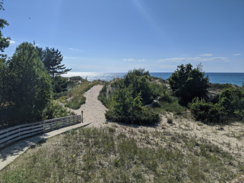 A sandy path leads to a beach, surrounded by greenery and trees, with the ocean visible in the background under a clear sky.