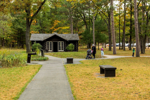 A path leads to a log cabin surrounded by trees, with people walking and a stroller in a park setting.
