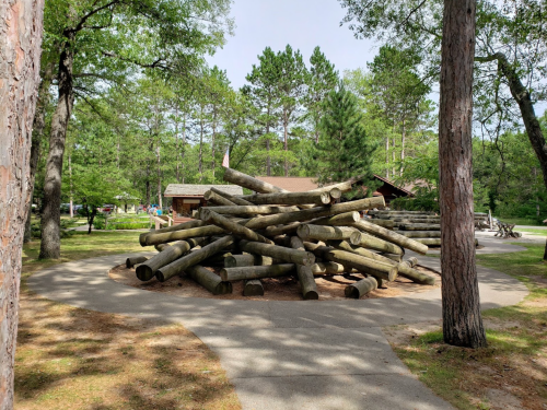 A pile of logs stacked in a park, with trees and a building in the background under a clear sky.