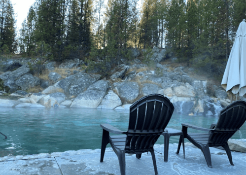 Two black lounge chairs by a steaming pool, surrounded by rocky terrain and tall trees in a serene outdoor setting.