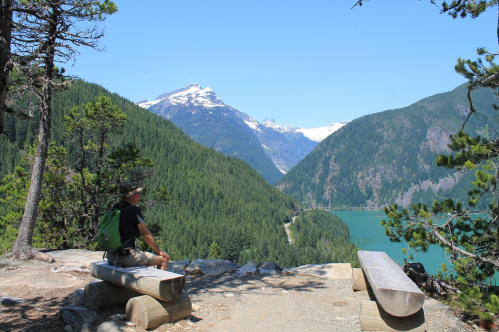 A hiker sits on a log overlooking a lush valley with mountains and a turquoise lake under a clear blue sky.