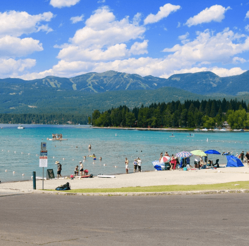 A sunny beach scene with people swimming, sunbathing, and enjoying the water, surrounded by mountains and trees.