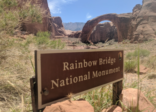 Sign for Rainbow Bridge National Monument with the iconic rock arch in the background under a clear blue sky.