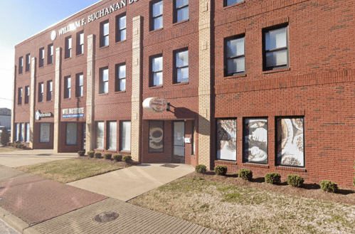 Exterior view of a brick building with multiple windows and signage, featuring a landscaped sidewalk in front.