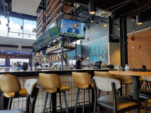 A modern bar with high stools, multiple TVs showing news, and a neon sign reading "101 TAPS" in a stylish setting.