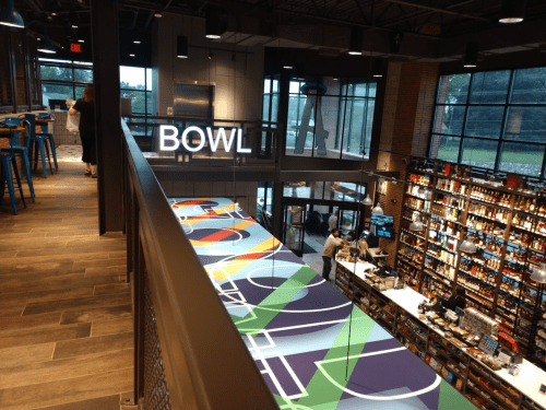 Interior of a modern bowling alley with a vibrant bar area and shelves filled with drinks. Large "BOWL" sign visible.