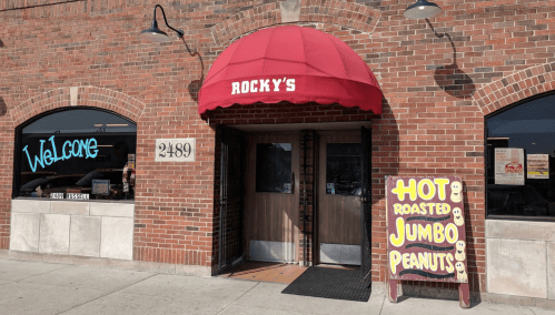 Entrance of Rocky's with a red awning, welcoming sign, and a board advertising hot roasted jumbo peanuts.