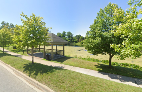 A gazebo beside a pond surrounded by green trees and grass on a sunny day.