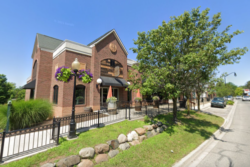 A brick building with large windows, surrounded by greenery and decorative flowers, on a sunny street.