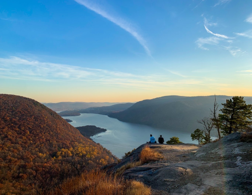 Two people sit on a rocky ledge overlooking a river and mountains at sunset, surrounded by autumn foliage.