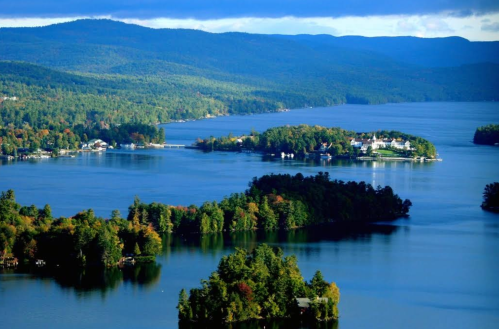 A scenic view of a lake surrounded by lush green forests and distant mountains under a partly cloudy sky.