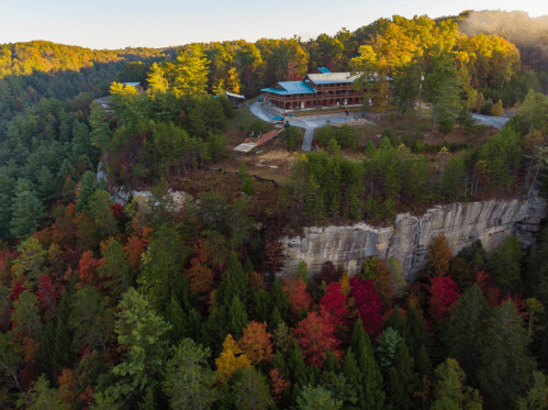 Aerial view of a lodge on a cliff surrounded by vibrant autumn foliage and lush green trees.
