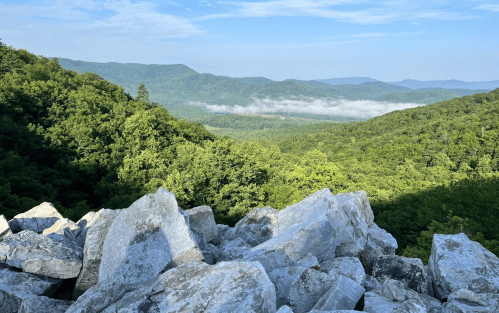A scenic view of lush green mountains and valleys under a clear blue sky, with large rocks in the foreground.