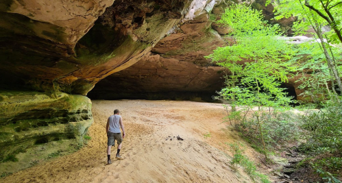 A person walks on sandy ground inside a large rock formation, surrounded by greenery and trees.