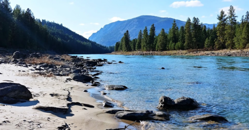 A serene river flows between mountains, with sandy shores and scattered rocks under a clear blue sky.