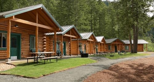 A row of rustic log cabins surrounded by trees, with picnic tables and a gravel path in a serene outdoor setting.