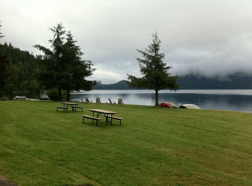 A serene lakeside view with picnic tables, trees, and boats on calm water under a cloudy sky.