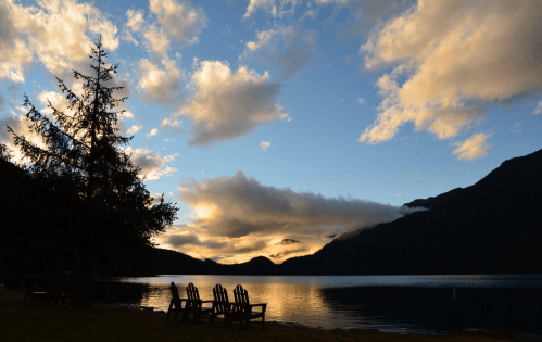 A serene lake at sunset, with silhouetted chairs and mountains under a colorful sky filled with clouds.