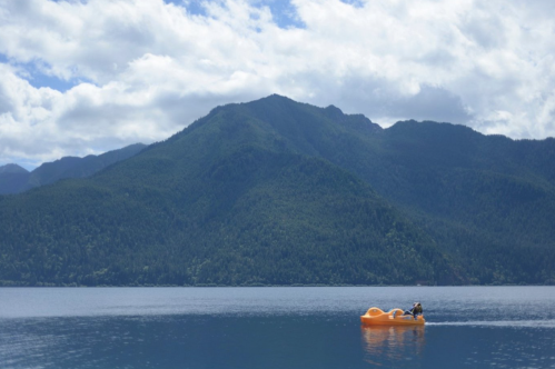 A person paddles an orange kayak on a calm lake, surrounded by lush green mountains under a partly cloudy sky.