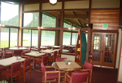 Cozy restaurant interior with wooden accents, tables, and large windows overlooking a scenic lake and forest.