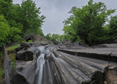 A rocky landscape with layered formations, surrounded by lush green trees under a cloudy sky.