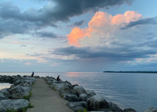 A serene lakeside scene with two people on a rocky pier, under a colorful sky with clouds at sunset.