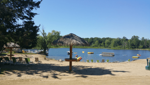 A sandy beach by a calm lake, featuring a thatched umbrella and colorful inflatable toys in the water.