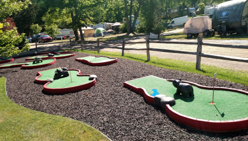 A mini-golf course with green turf and red borders, surrounded by trees and camping trailers in the background.