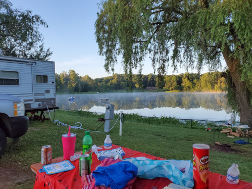 A serene lakeside view at dawn, with mist over the water and a campsite setup featuring snacks and drinks.