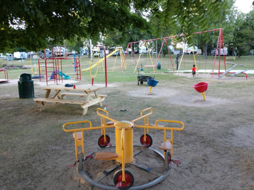 A playground with swings, a merry-go-round, and picnic tables, surrounded by trees and grassy areas.