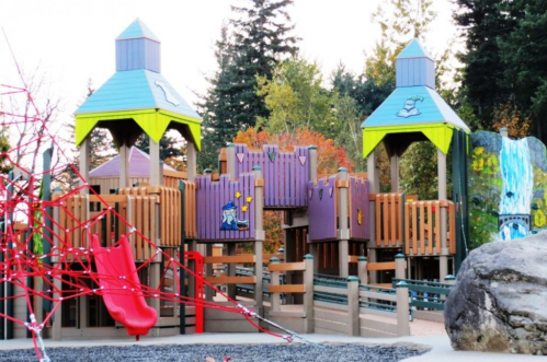 Colorful playground structure with slides, climbing features, and a rope net, surrounded by trees and rocks.