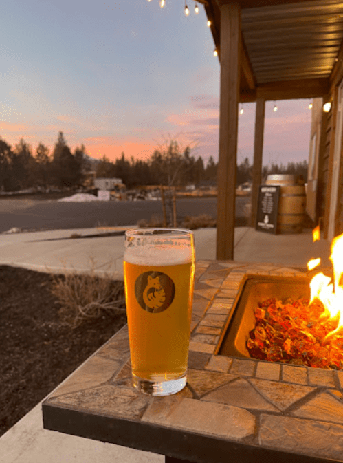 A glass of beer sits on a stone table next to a fire pit, with a sunset and outdoor scenery in the background.