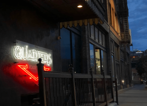 Neon sign for "Quarters" illuminated on a building exterior at dusk, with a quiet street in the foreground.