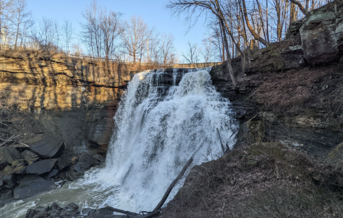 A cascading waterfall flows over rocky cliffs, surrounded by bare trees and a clear blue sky.