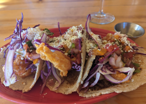 Three tacos filled with fried fish, cabbage, salsa, and cheese on a red plate, with a small bowl on the side.