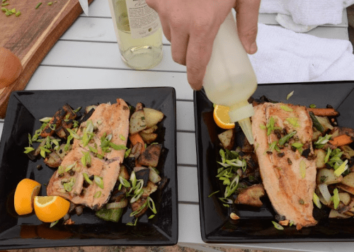 Two black plates with salmon fillets, roasted vegetables, and a hand drizzling sauce over the dish.