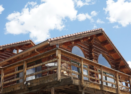 A wooden log cabin with large arched windows and a balcony, set against a blue sky with fluffy clouds.