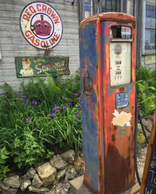 Vintage gas pump with peeling paint, surrounded by flowers and an old "Red Crown Gasoline" sign in the background.
