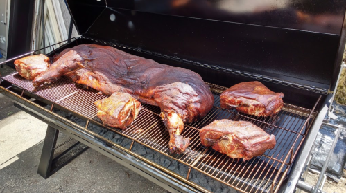 A large smoked pig and several smaller cuts of meat on a grill, showcasing a rich, dark, caramelized exterior.