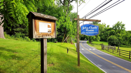 Sign for John Chads' House with historical information, set along a grassy roadside with trees in the background.