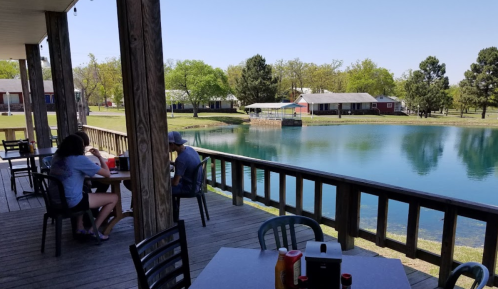 A scenic view of a deck by a pond, with people dining and trees in the background on a sunny day.