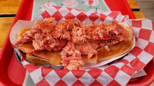 A lobster roll overflowing with lobster meat, served on a toasted bun and garnished with paprika, on a red-checkered tray.