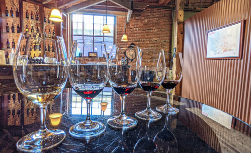 A row of wine glasses with varying levels of red and white wine, set against a rustic brick wall and wooden decor.