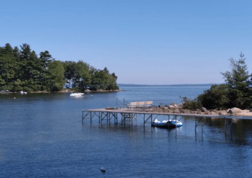 A calm lake scene featuring a wooden dock, a small boat, and lush green trees under a clear blue sky.