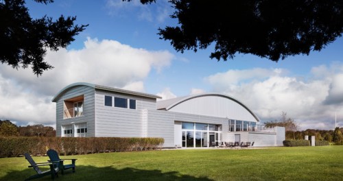 A modern, silver-clad building with large windows, set in a green landscape under a partly cloudy sky.