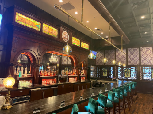 A stylish bar with stained glass, wooden accents, and teal seating, featuring shelves of colorful bottles.